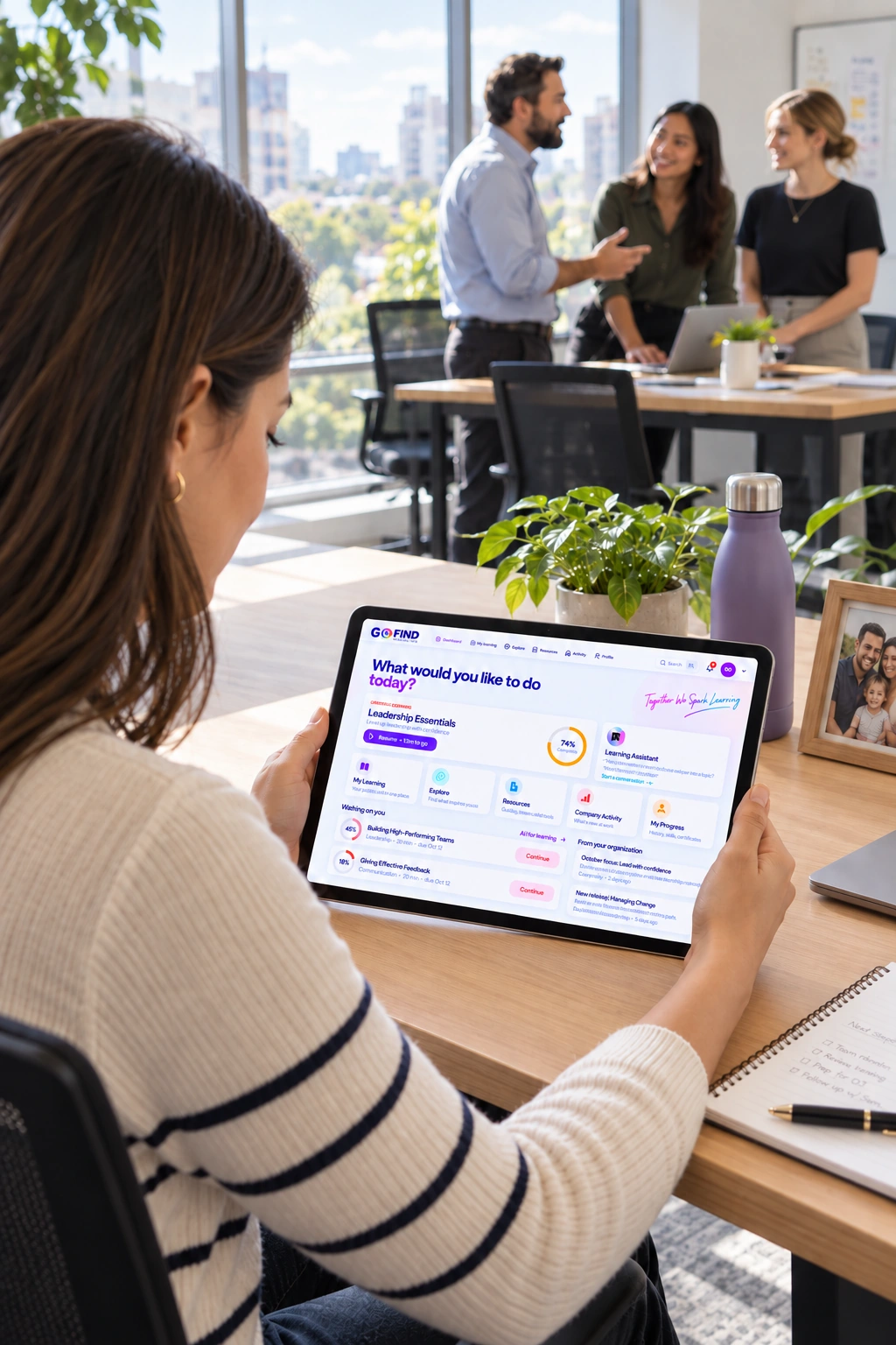 A woman using the Go Find Learning platform on a tablet with colleagues talking in the office behind her